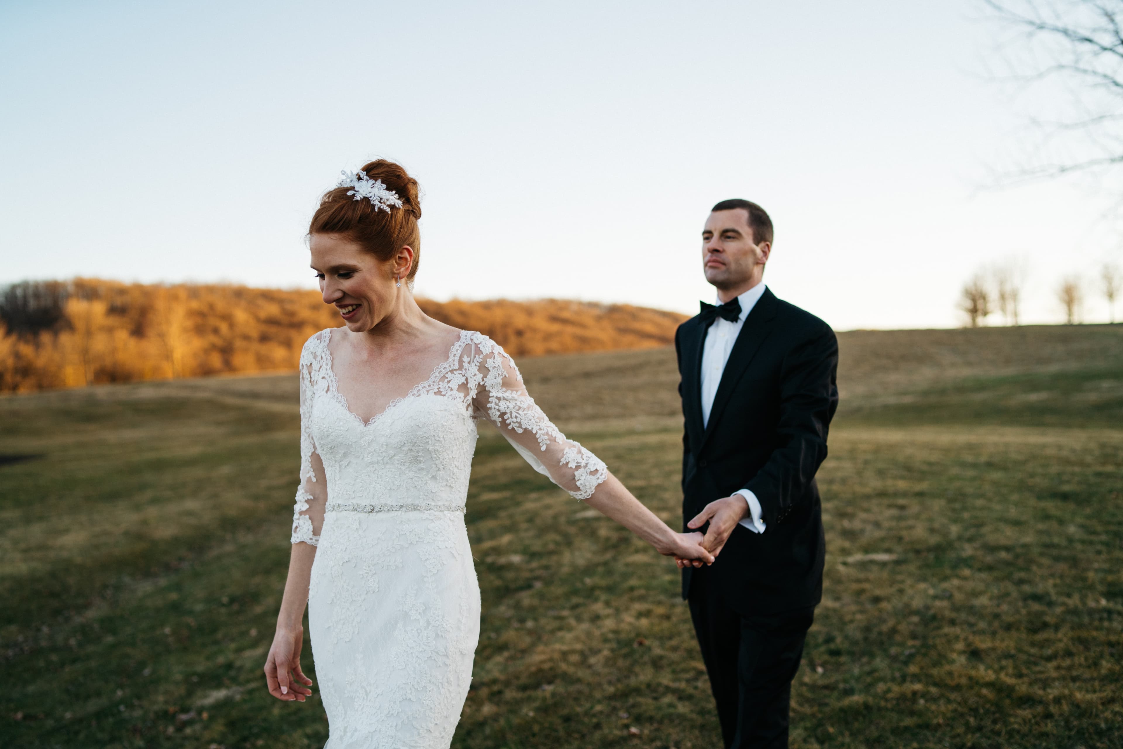 Elegant wedding couple in formal attire walking through autumn field at sunset